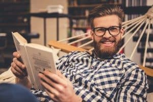 Handsome bearded young man is reading a book, looking at camera and smiling while lying in the hammock in modern library